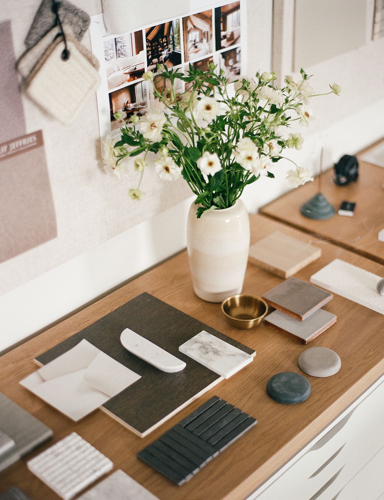Design desk holding white flowers in a ceramic vase, marble and wood tile samples, a brass bowl, and a pinned fabric mood board.