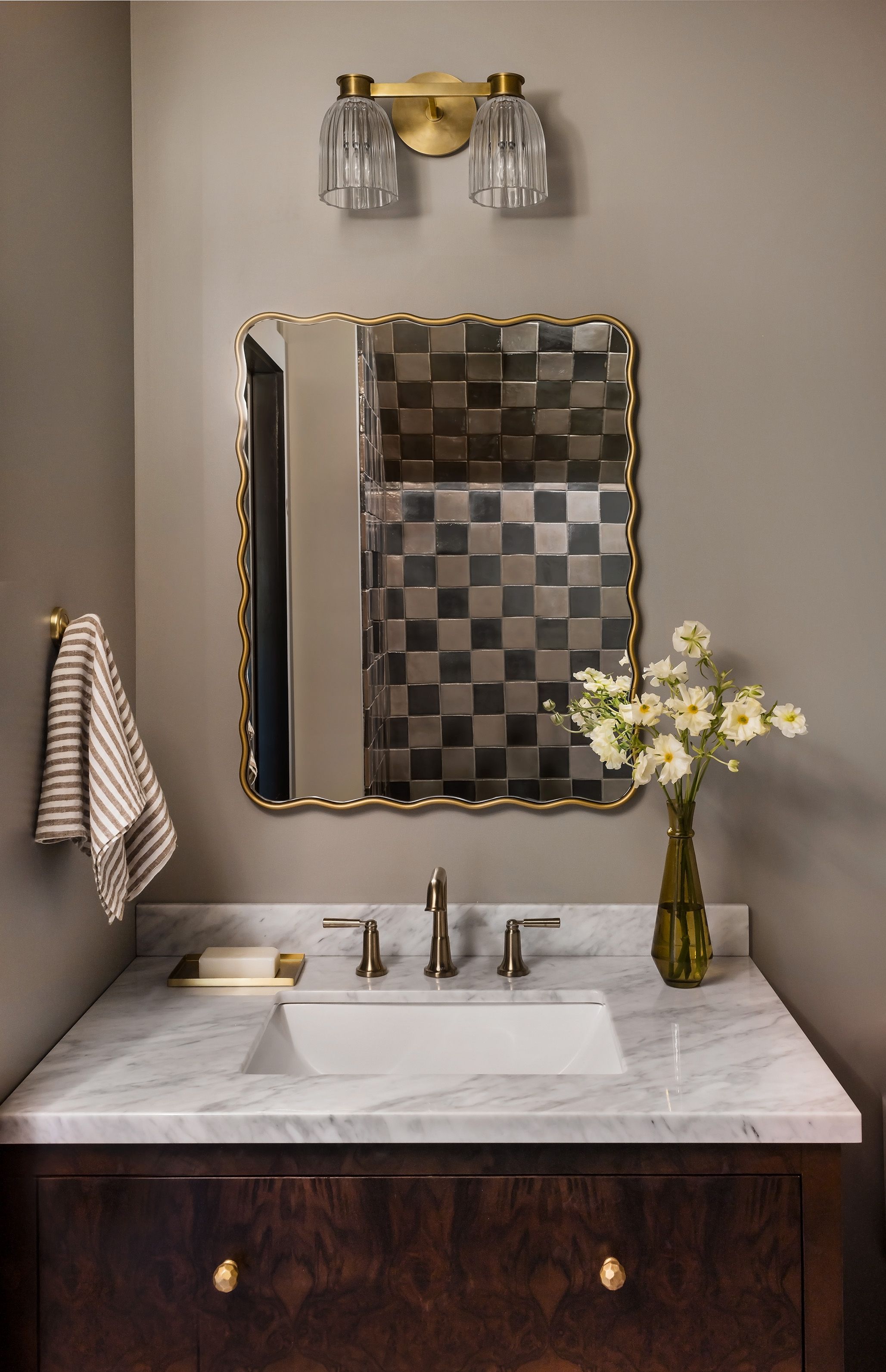 A marble bathroom vanity reflects a black-and-tan checkerboard tile shower in its scalloped brass mirror, beside a striped linen towel.
