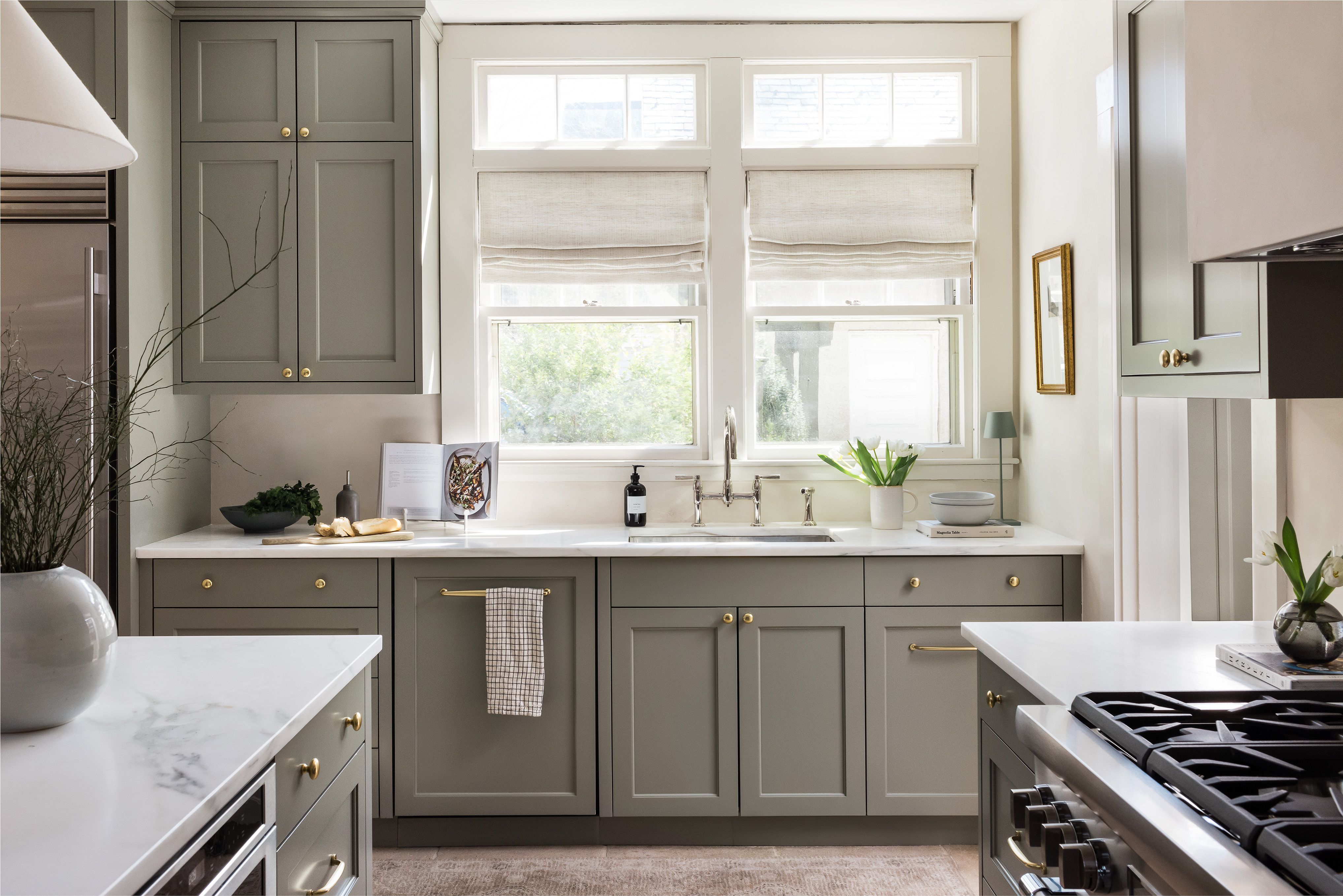A kitchen with sage cabinetry, brass hardware, linen roman shades over the sink window, and a small gold-framed print on the wall.