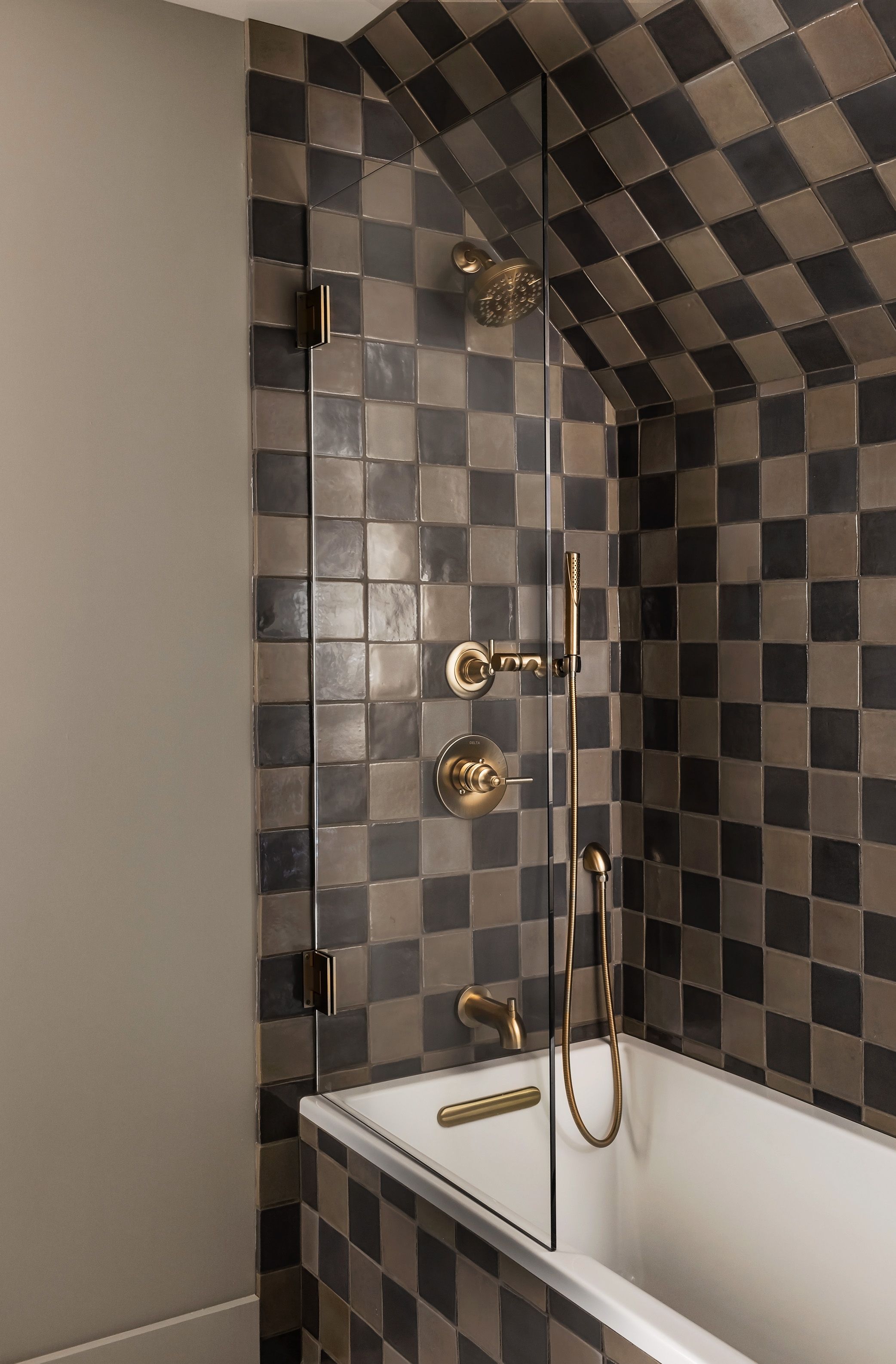 A bathtub alcove is wrapped in brown-and-tan checkerboard tile, with brass fixtures and a handheld shower on a frameless glass panel.