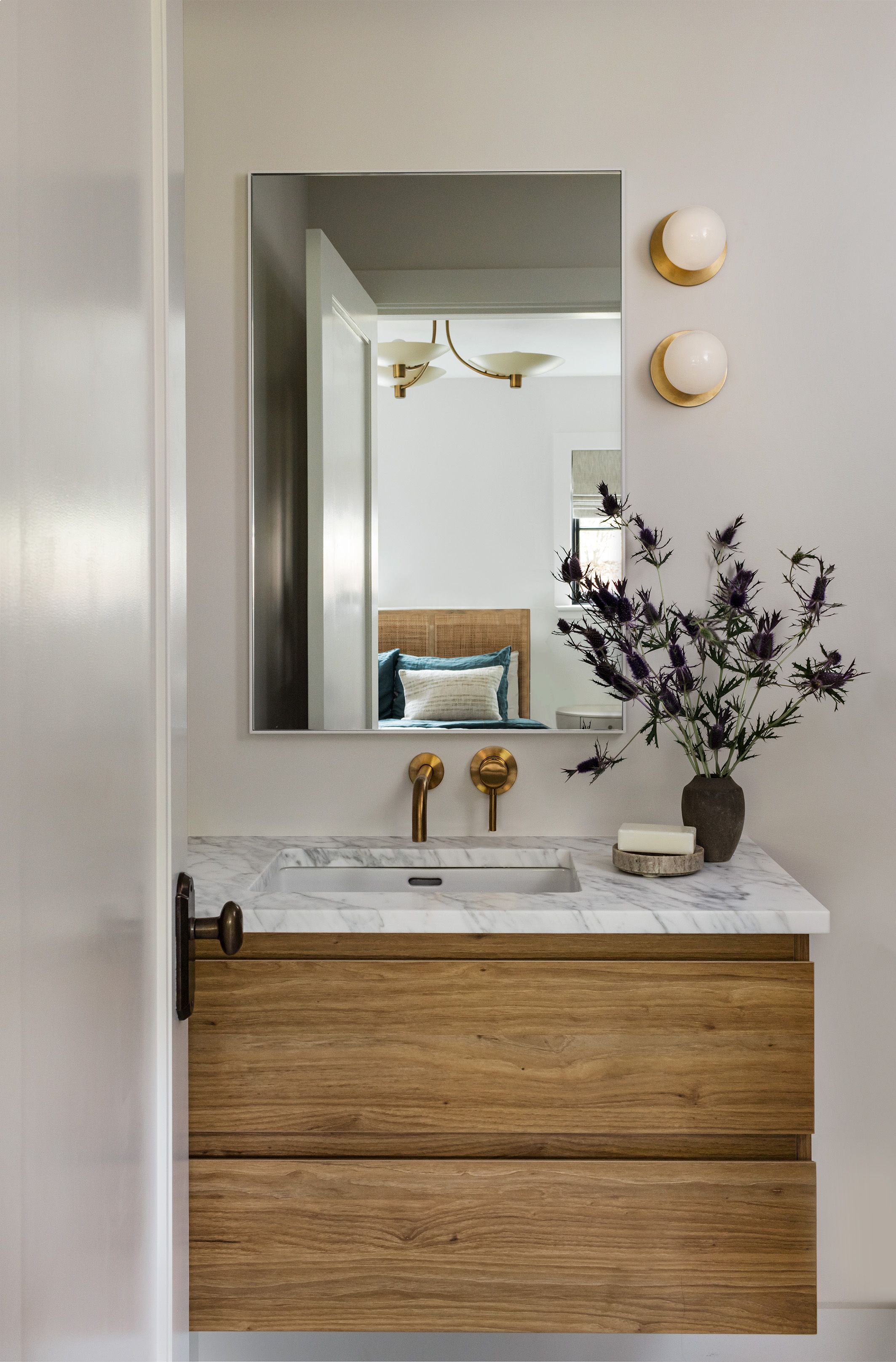 A powder room vanity with a black stone counter and terrazzo-tile backsplash reflects a bedroom in its arched mirror flanked by brass sconces.