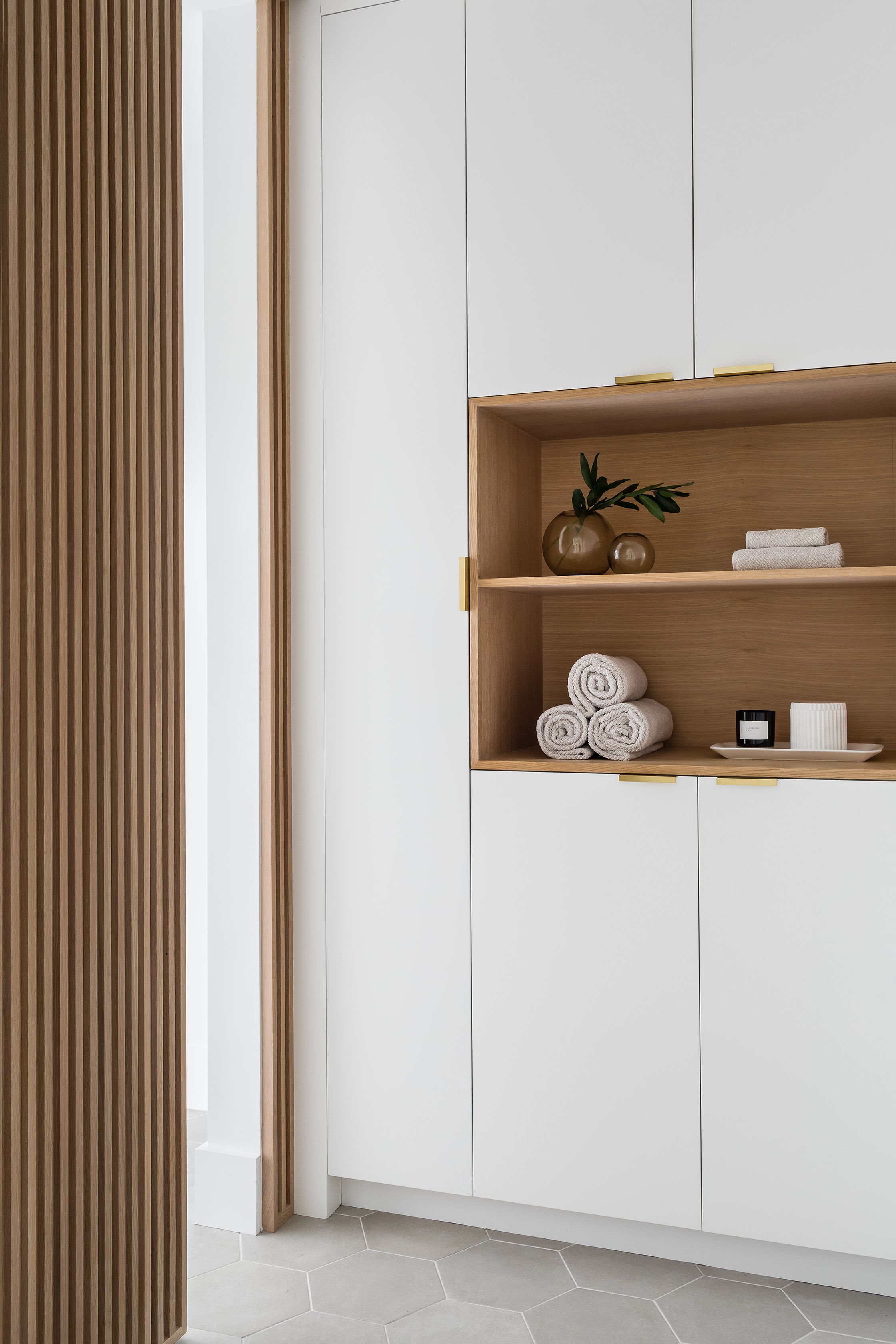 Built-in linen cabinetry with a wood slat screen and an open oak shelf holding rolled towels, bud vases, and a candle.