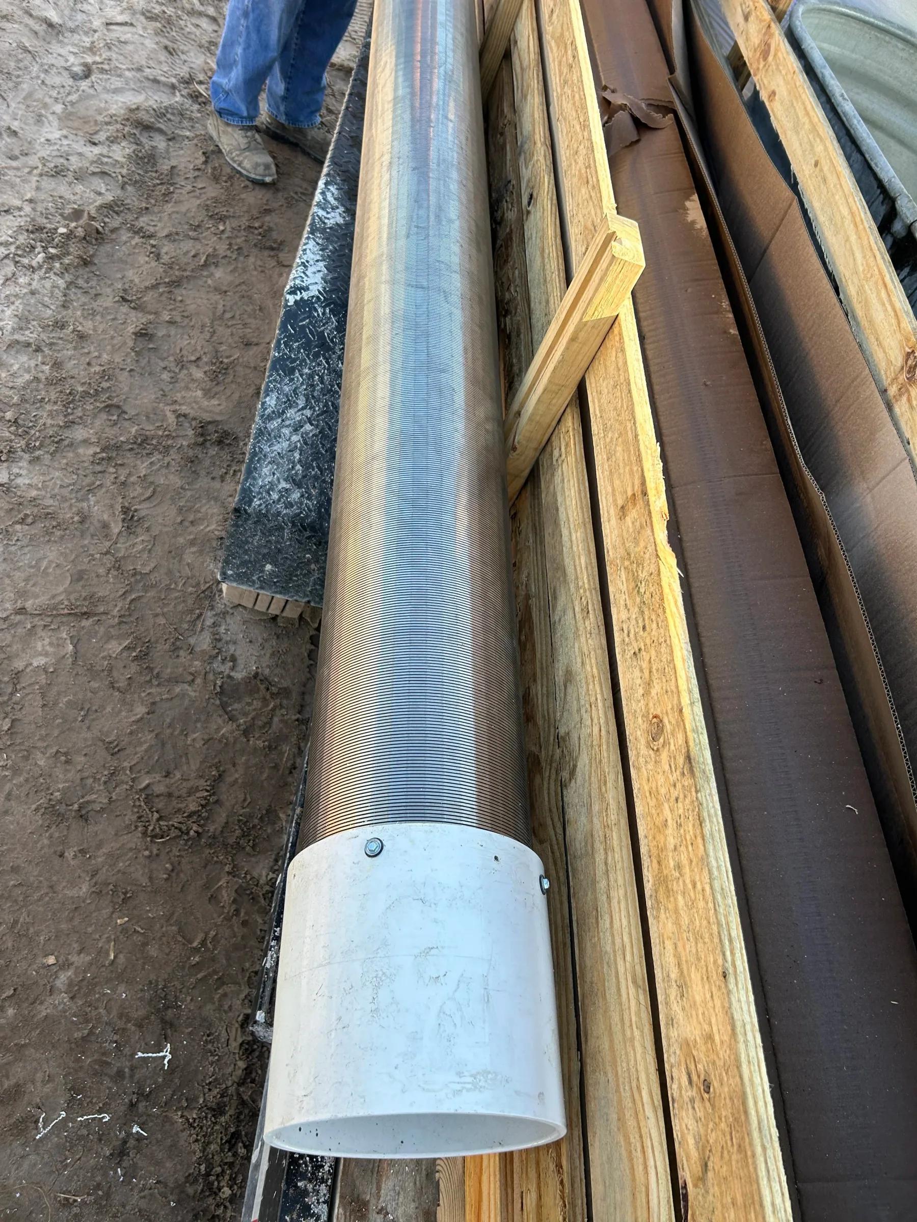 A length of metal well pipe with a white fitting, laid on boards on site
