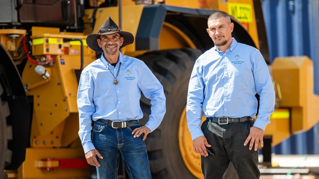 Two Indigenous men standing in front of mining machinery