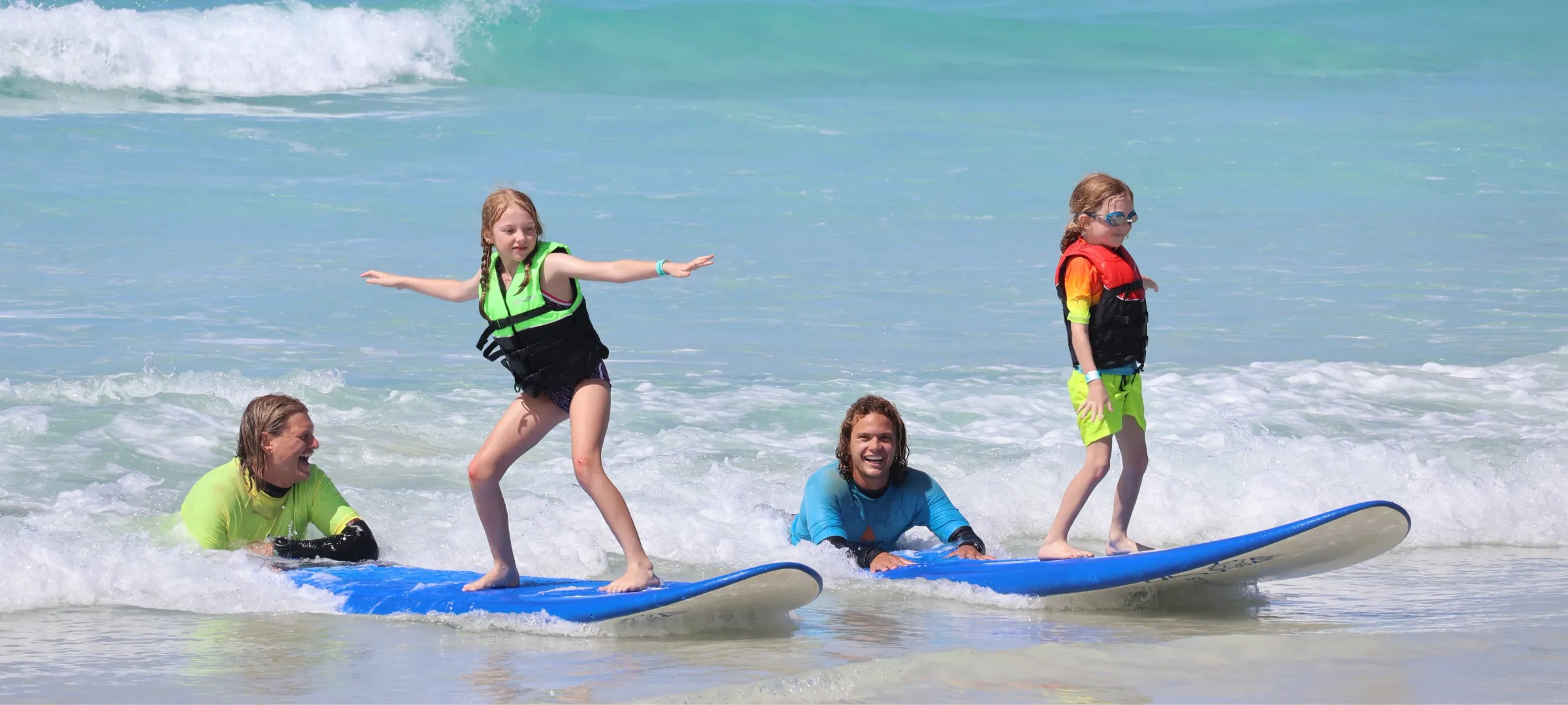 Two kids on surfboards in the ocean with helpers