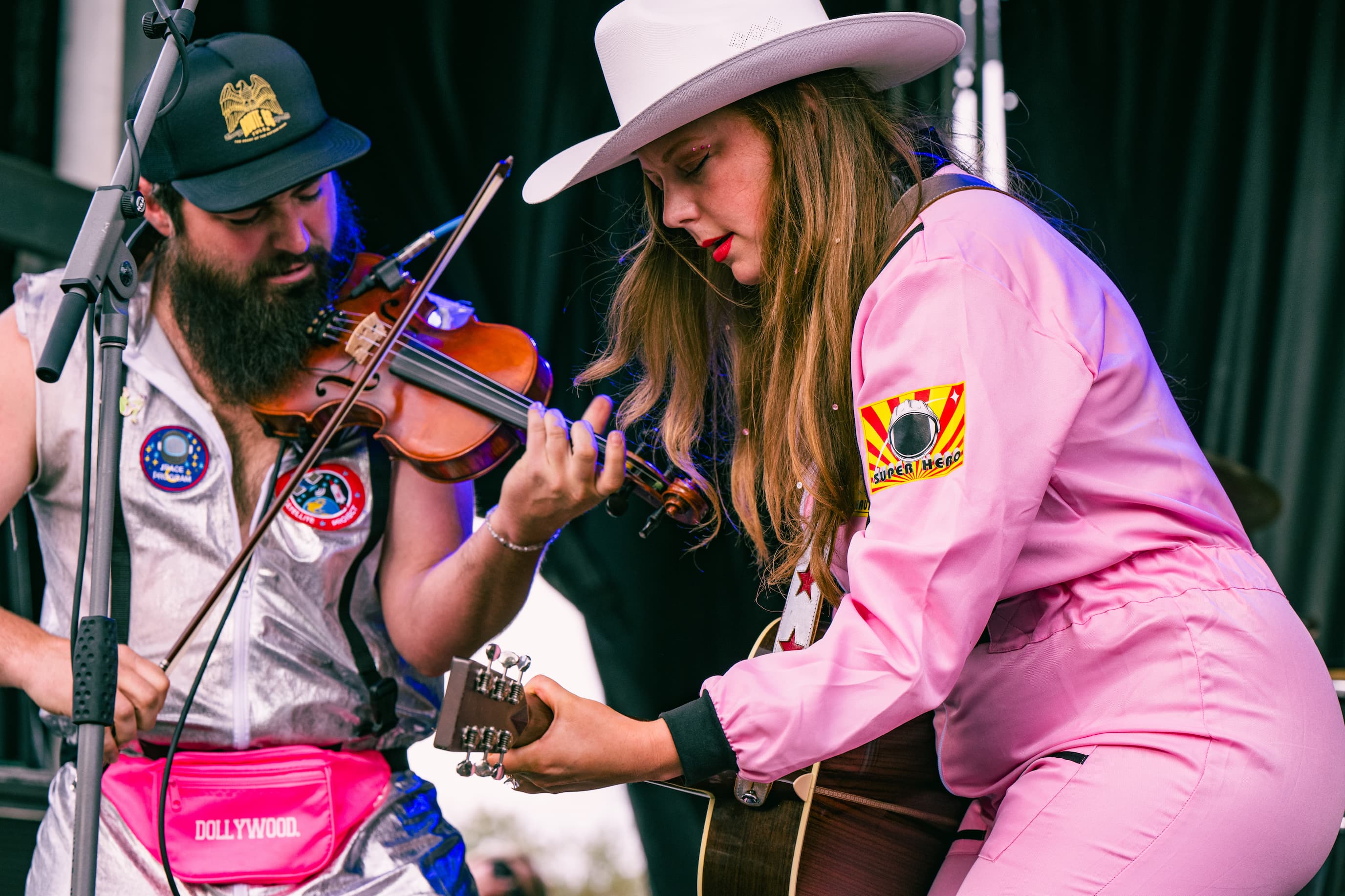 Kaitlin Butts in a pink spacesuit and cowboy plays guitar next to fiddle player in a silver space suit