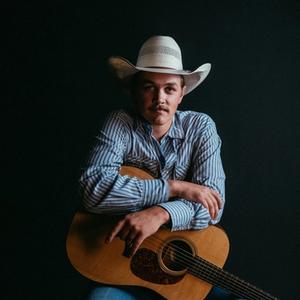 Zach Top in blue striped shirt and white cowboy hat starting straight into the camera while leaning on a guitar in his lap.
