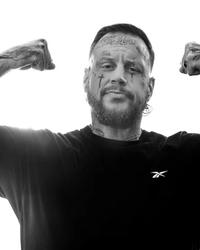 Jelly Roll flexes his arms while wearing a black reebok t-shirt, sporting his tattoos in a black and white photo.