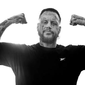 Jelly Roll flexes his arms while wearing a black reebok t-shirt, sporting his tattoos in a black and white photo.
