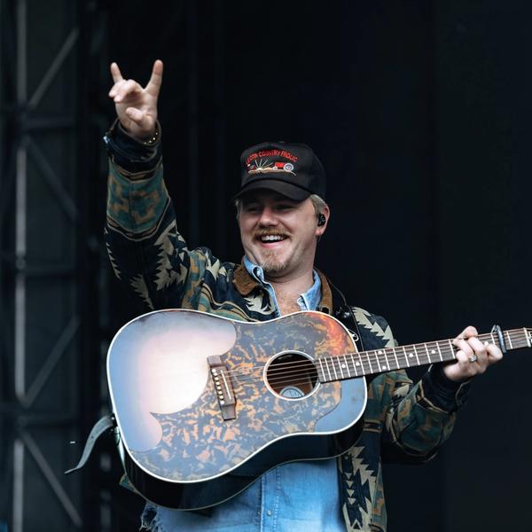 Ernest give the rock sign and laughing while holding a guitar and wearing a denim shirt, wool pattern jacket and black Leota Country Frolic trucker cap on stage at Two Step Inn 2024.
