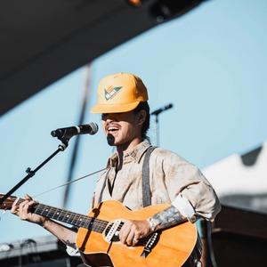Nat Myers wearing a yellow hat and tan shirt, singing into a mic while playing guitar.