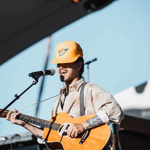 Nat Myers wearing a yellow hat and tan shirt, singing into a mic while playing guitar.