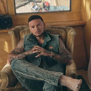 Kane Brown sat in a coach in front of a window, in a denim vest jumpsuit.