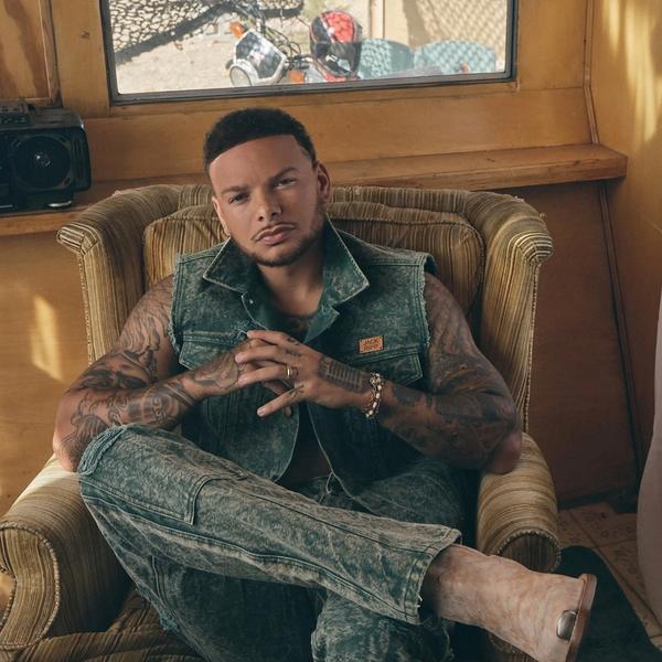 Kane Brown sat in a coach in front of a window, in a denim vest jumpsuit.