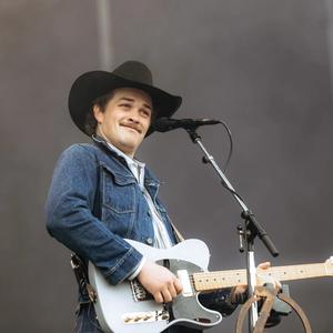 Zach Top stares into the crowd with a smirk on his face while playing an electric guitar in a denim jacket, blue striped shirt and black cowboy hat, with blue jeans on.