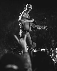 Riley Green on a stage above a crowd of people, playing a guitar while wearing jeans, a shirt and a trucker cap. Photographed in black and white.