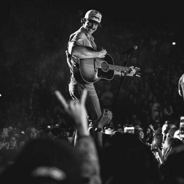 Riley Green on a stage above a crowd of people, playing a guitar while wearing jeans, a shirt and a trucker cap. Photographed in black and white.