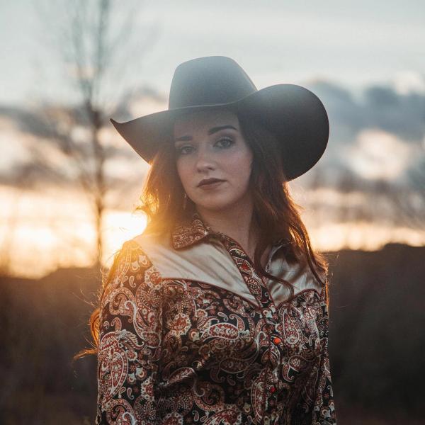 Noeline Hoffman, in paisley western shirt and black Stetson hat, looks into the camera in front of a out of focus, sunset background.
