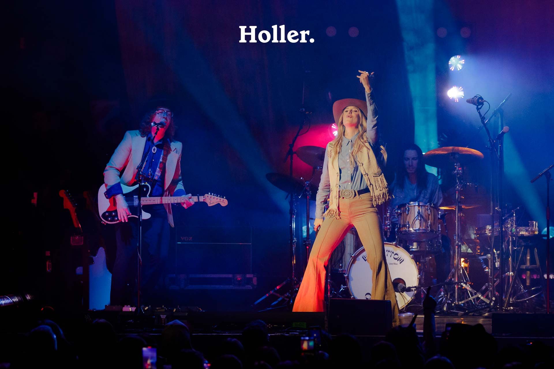 <p>Lainey Wilson points into the sky with a microphone in her hand, while wearing a beige waistcoat, denim shirt, brown bell bottom jeans and a brown hat, with a guitarist playing guitar to the right of her and a drummer playing drums to her left.</p>