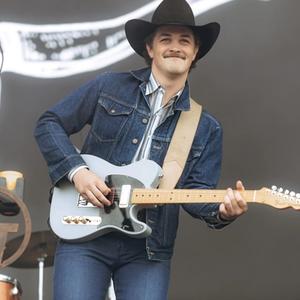 Zach Top playing an electric guitar in a denim jacket, blue striped shirt and black cowboy hat, with blue jeans on. A drummer sits behind him. 