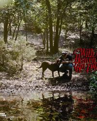 A man kneels and pets his dog near to a river amongst trees within a forest.
