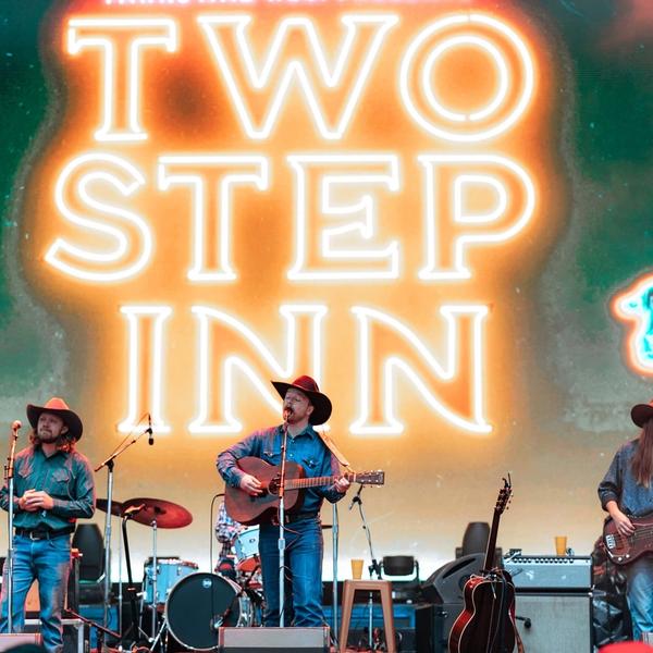 Colter Wall in front of a blue and orange Two Step Inn neon sign on stage at Two Step Inn, wearing a black cowboy hat, denim jacket while singing into a microphone and playing a guitar next to two of his band members, one playing a bass guitar. 
