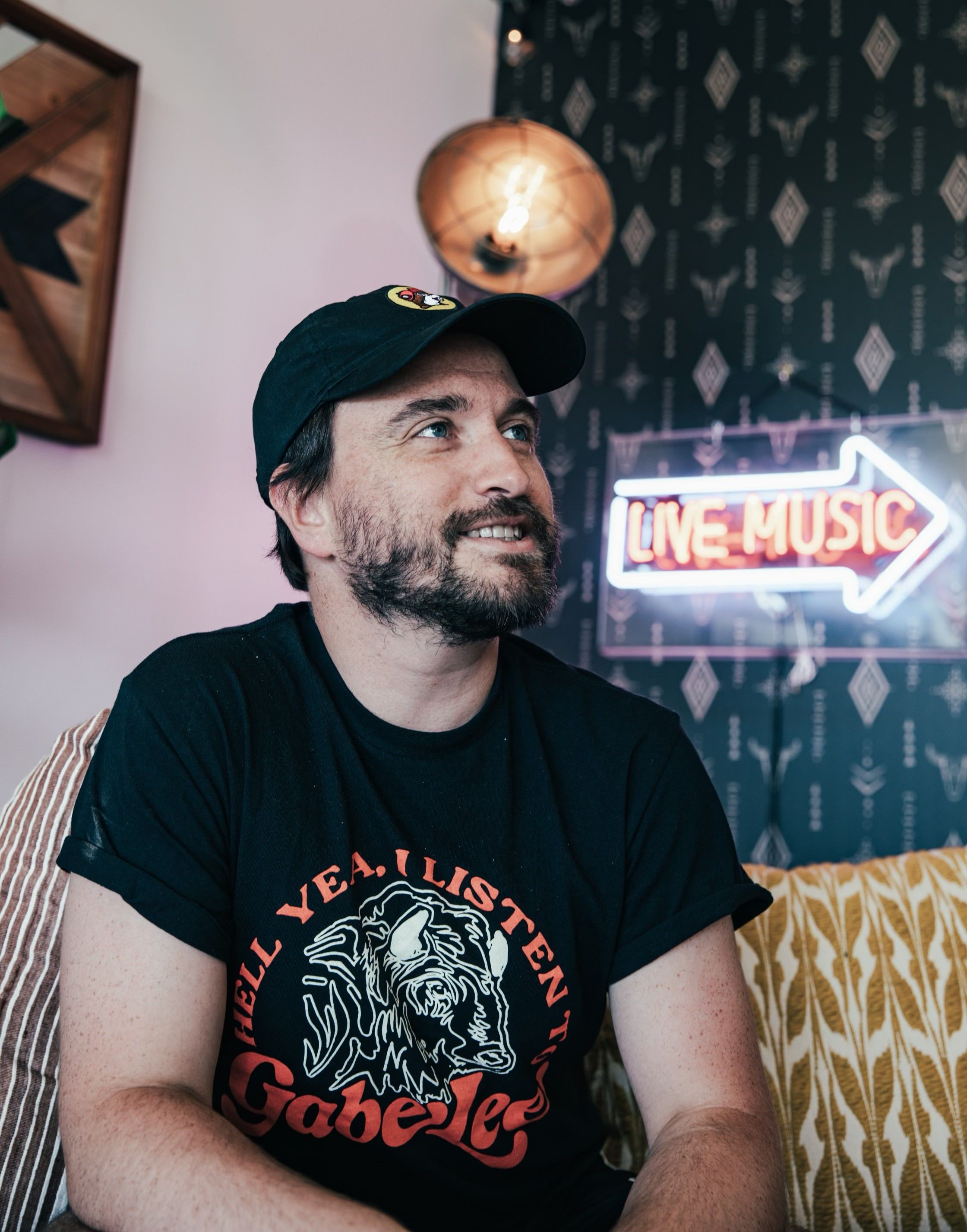 Ross Jones in a cap sat on a sofa looking up to the right, with a 'live music' in an arrow sign to the right of them.
