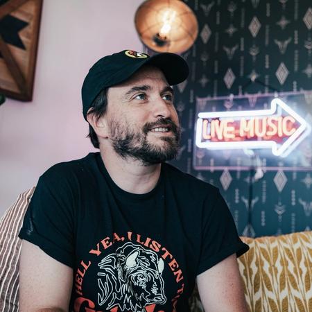 Ross Jones in a cap sat on a sofa looking up to the right, with a 'live music' in an arrow sign to the right of them.