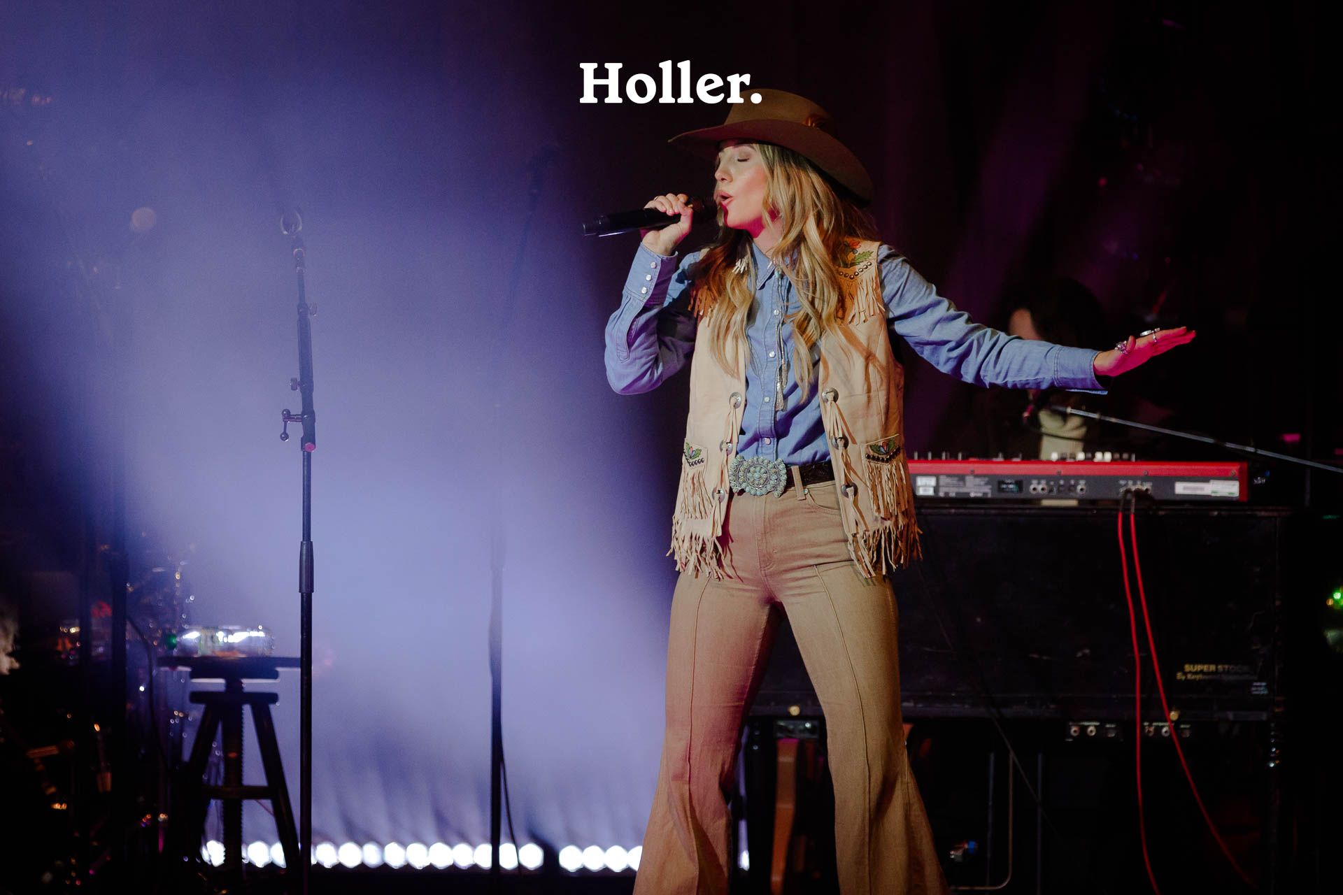 <p>Lainey Wilson sings into a microphone while wearing a beige waistcoat, denim shirt, brown bell bottom jeans and a brown hat, with a keyboard behind her.</p>