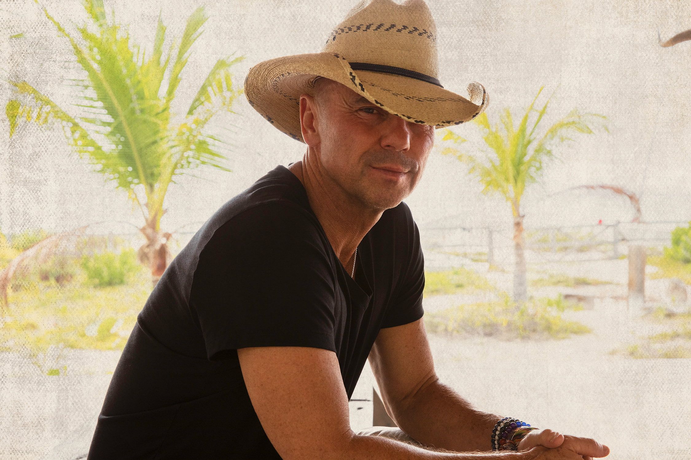 Kenny Chesney wearing a black t-shirt and straw cowboy hat in front of a transparent material backdrop with palm trees in the distance.