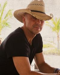 Kenny Chesney wearing a black t-shirt and straw cowboy hat in front of a transparent material backdrop with palm trees in the distance.