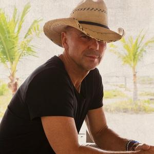 Kenny Chesney wearing a black t-shirt and straw cowboy hat in front of a transparent material backdrop with palm trees in the distance.