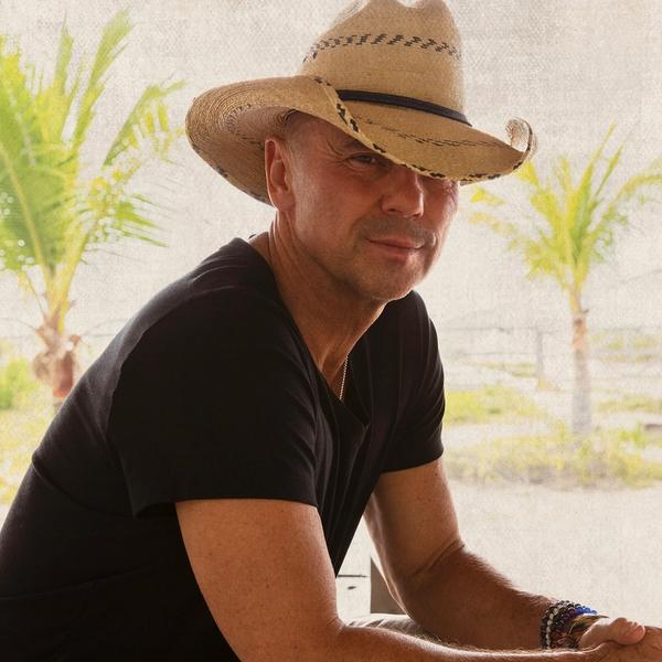 Kenny Chesney wearing a black t-shirt and straw cowboy hat in front of a transparent material backdrop with palm trees in the distance.