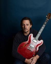 Sturgill Simpson sat holding a red guitar and wearing a black longsleeve top.