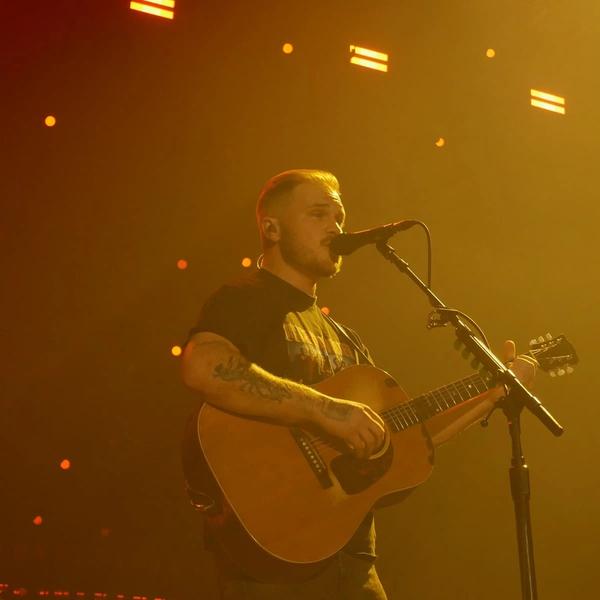 Zach Bryan singing into a microphone and playing a guitar in a black t-shirt infront of a yellow-lit backdrop.