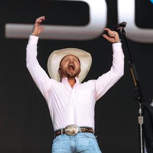 Cody Johnson shouting with his arms up in the air in front of a black backdrop while wearing a white cowboy hat, pink shirt and denim jeans with a brown belt and silver buckle.