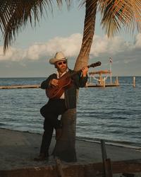 Ernest leaning against a palm tree, wearing a double denim suit and white cowboy hat, stood on a beach in front of the ocean on an evening.