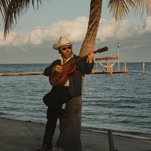 Ernest leaning against a palm tree, wearing a double denim suit and white cowboy hat, stood on a beach in front of the ocean on an evening.