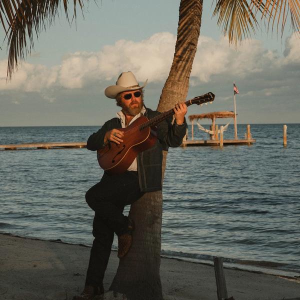 Ernest leaning against a palm tree, wearing a double denim suit and white cowboy hat, stood on a beach in front of the ocean on an evening.