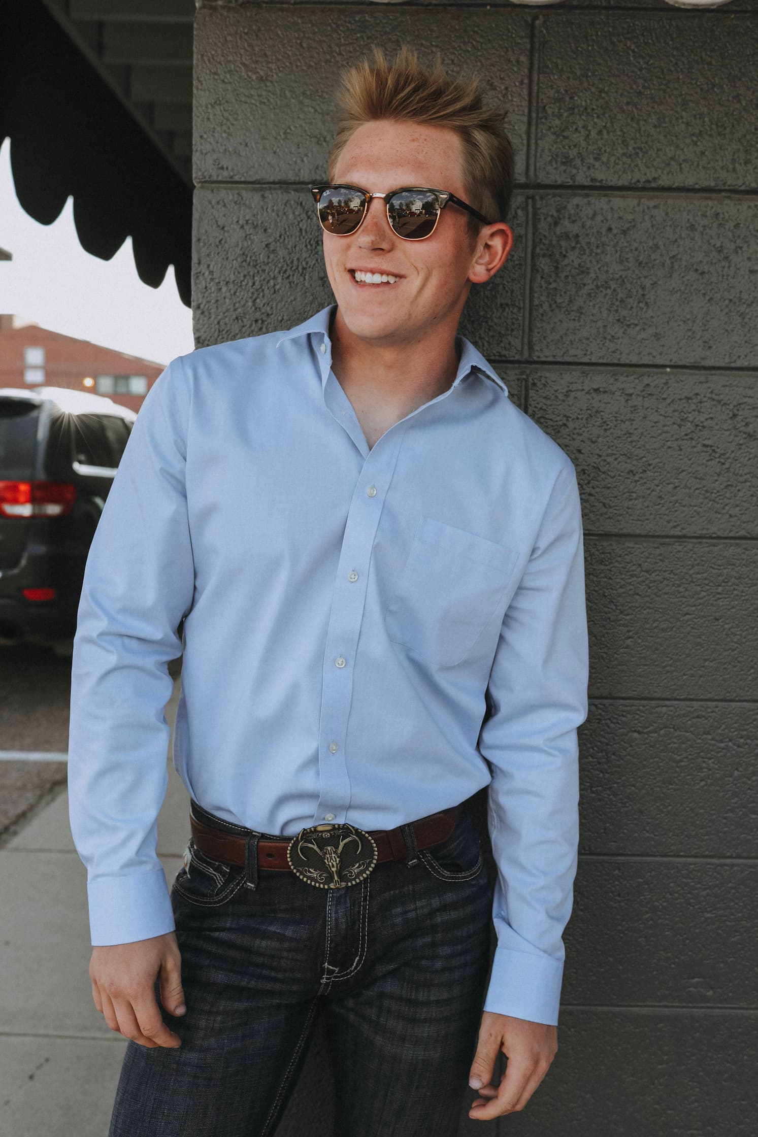 A portrait of Kyle Amaral wearing sunglasses, blue jeans and a brown bronco belt in front of a grey wall.