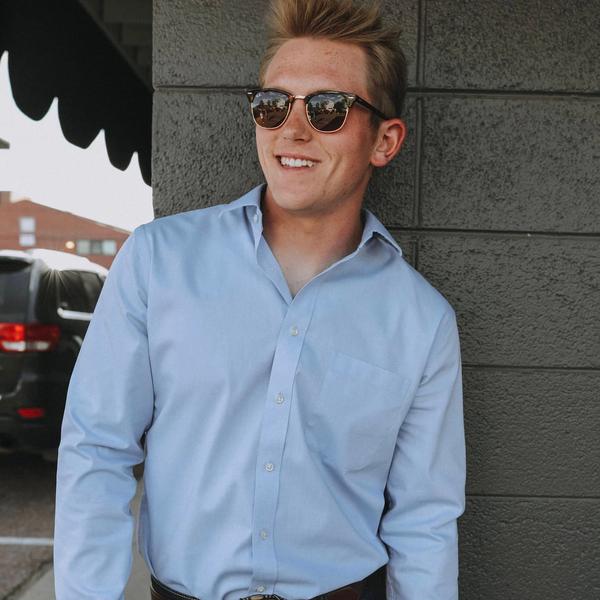 A portrait of Kyle Amaral wearing sunglasses, blue jeans and a brown bronco belt in front of a grey wall.