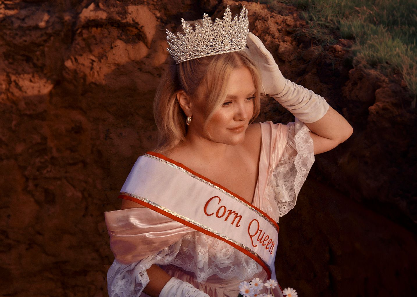 Hailey Whitters wearing a tiara, debutant white dress and gloves while holding flowers. She's wearing 'Corn Queen' sash.
