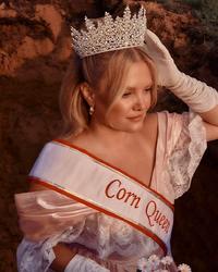 Hailey Whitters wearing a tiara, debutant white dress and gloves while holding flowers. She's wearing 'Corn Queen' sash.