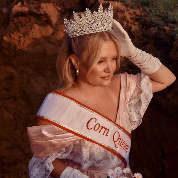 Hailey Whitters wearing a tiara, debutant white dress and gloves while holding flowers. She's wearing 'Corn Queen' sash.