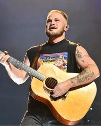 Zach Bryan On Stage Playing A Guitar Wearing A Black Vest