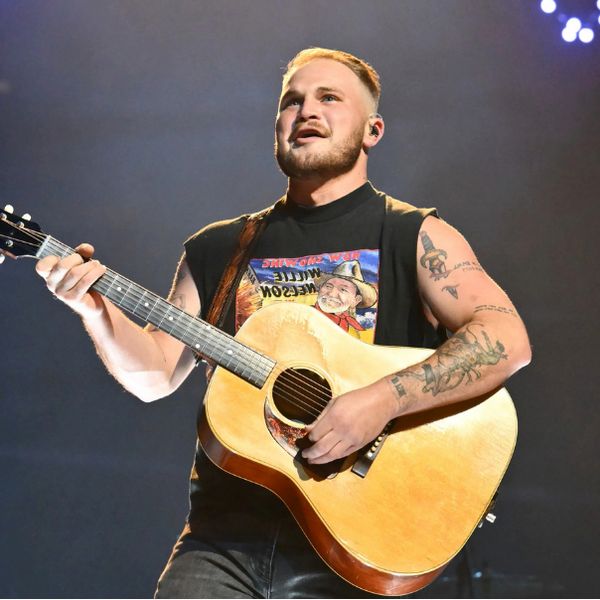 Zach Bryan On Stage Playing A Guitar Wearing A Black Vest
