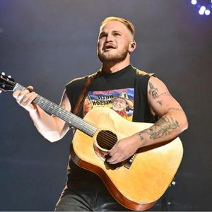 Zach Bryan On Stage Playing A Guitar Wearing A Black Vest