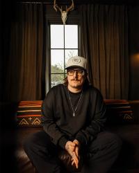 Hardy sat in a bedroom in front of a long narrow window, with a buck head hung above the him. He wears a white trucker cap, black longsleeve top and tanned sunglasses.