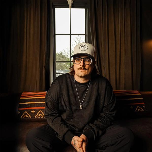 Hardy sat in a bedroom in front of a long narrow window, with a buck head hung above the him. He wears a white trucker cap, black longsleeve top and tanned sunglasses.