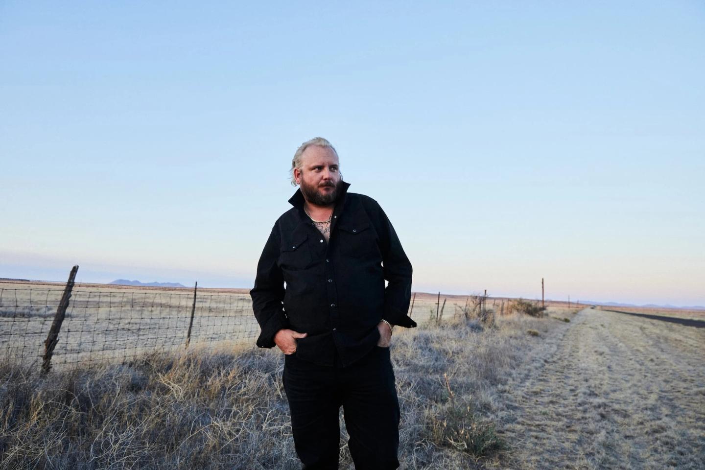 Paul Cauthen, in a field, standing in front of a wire fence, dressed in all black with a upturned collar on his black shirt with rolled-up sleeves.